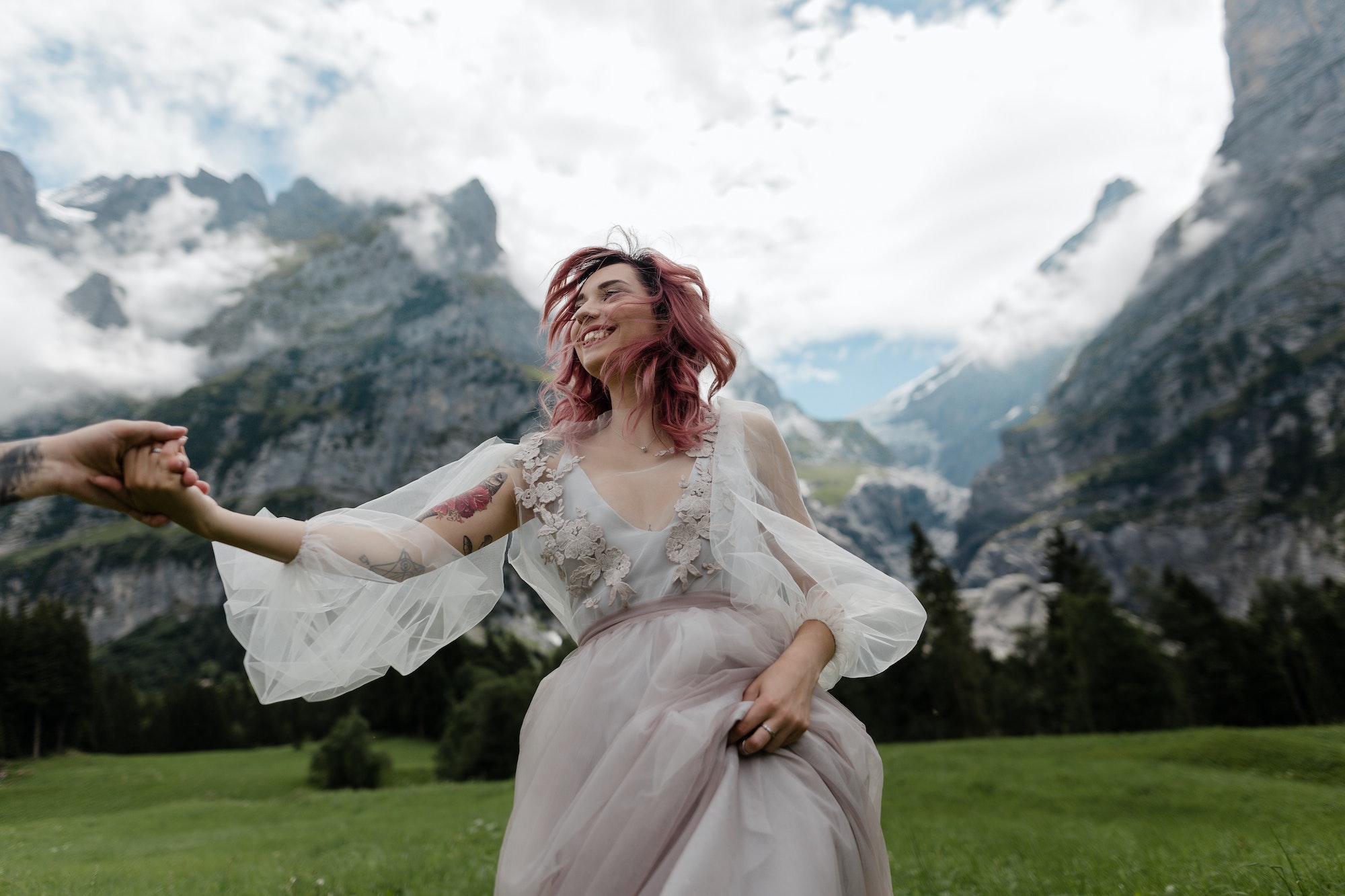 Happy Bride in Wedding Dress Holding Hands With Groom on Meadow With Mountains and Clouds in Alps