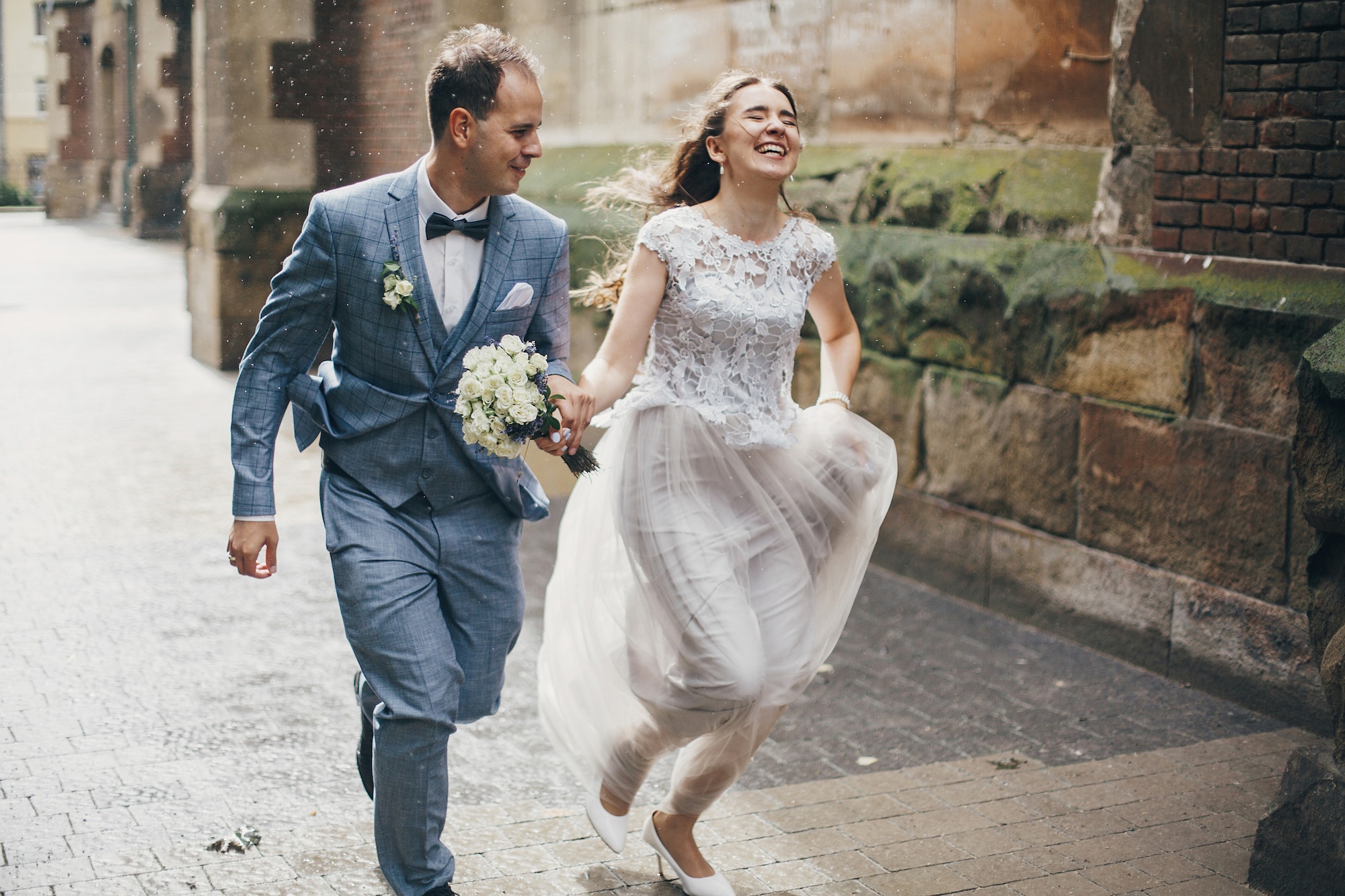 Stylish happy bride and groom running on background of old church in rainy street. Provence wedding