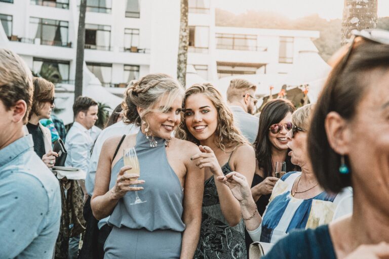 Twee lachende vrouwen in menigte van bruiloft, met drankjes in de hand - Jurk bruiloft gast | Volvo Trouwauto